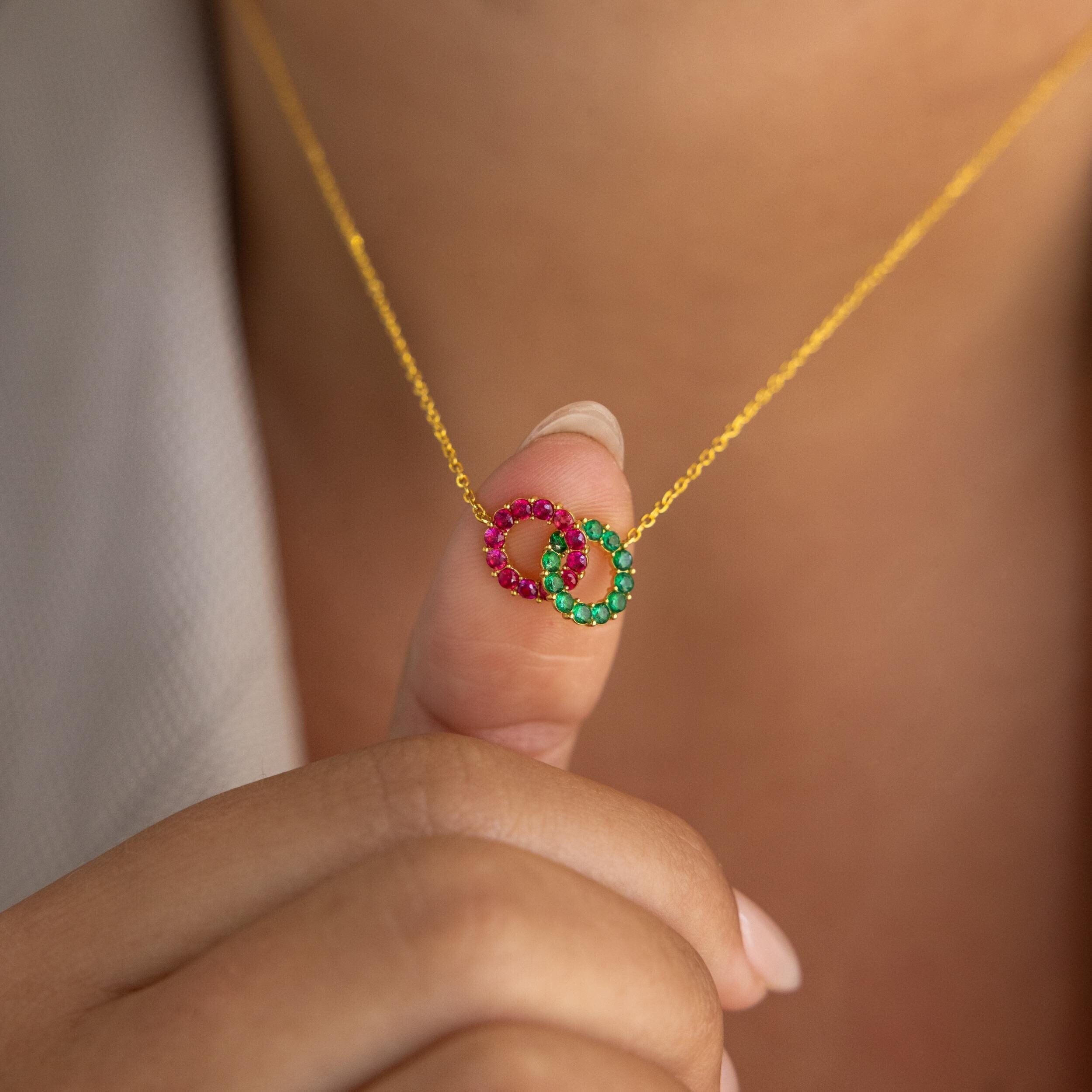 A close-up image of a hand holding a gold necklace with an interlocking duo birthstone charm in silver.